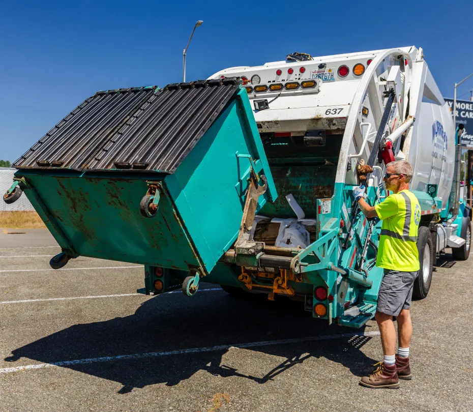 Worker operating mechanism to empty a green commercial dumpster into a white garbage truck.