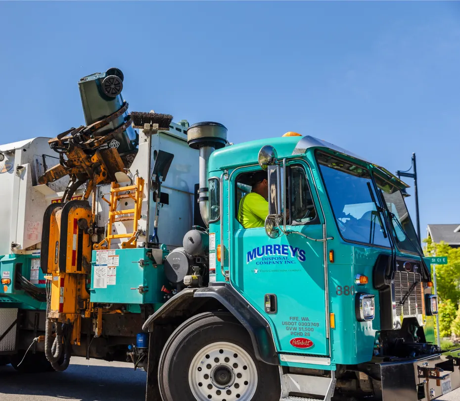 Garbage truck from Murrey's Disposal lifting a green curbside bin against a clear blue sky.