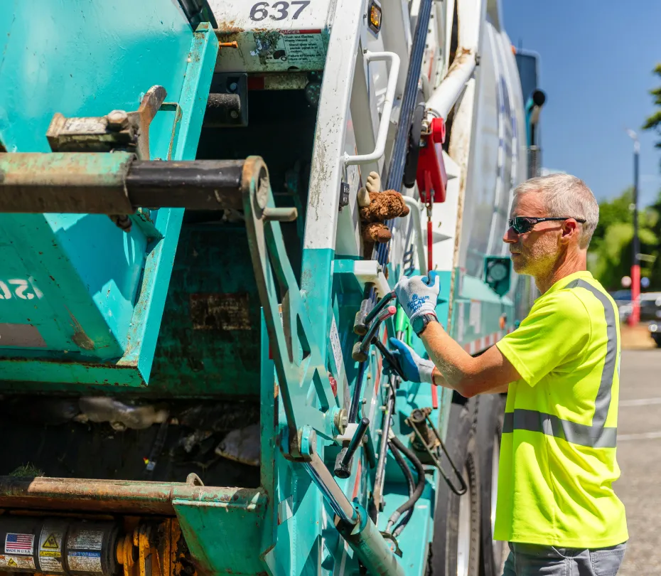 Worker in neon yellow safety shirt operating controls on a rear load garbage truck.