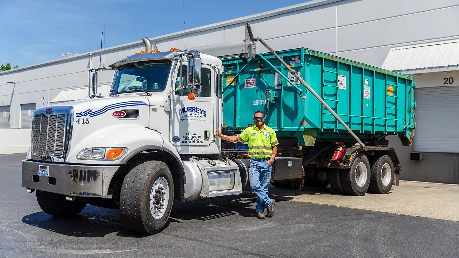 Man in yellow safety shirt standing beside white roll-off dumpster truck in an industrial area.