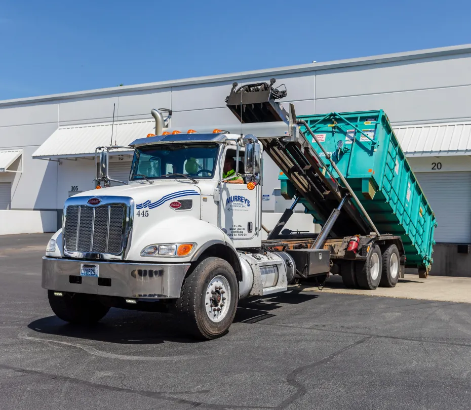 White roll-off truck lifting a large green dumpster near a commercial building's loading dock.
