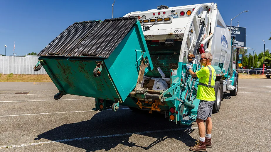 Worker in yellow safety shirt operates a truck lift to empty a large blue commercial dumpster.