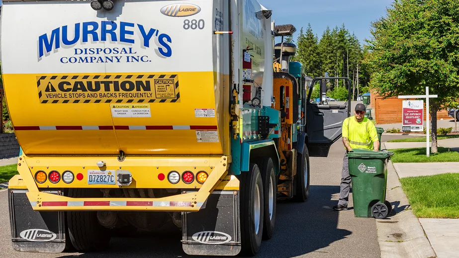 Worker in neon yellow shirt prepares green trash cart beside Murrey's Disposal garbage truck on a neighborhood street.