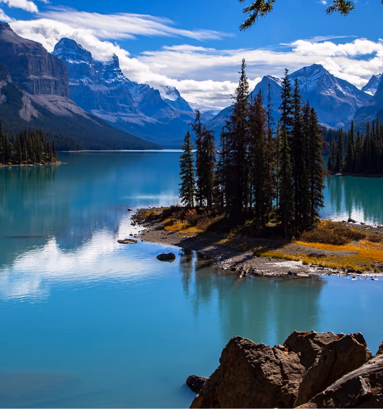 Turquoise lake surrounded by pine trees and rocky snow-capped mountains under a blue sky.