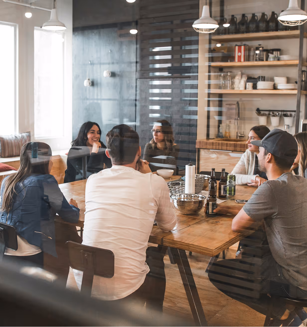 Group of six people sitting around a wooden table in a modern kitchen or office space.