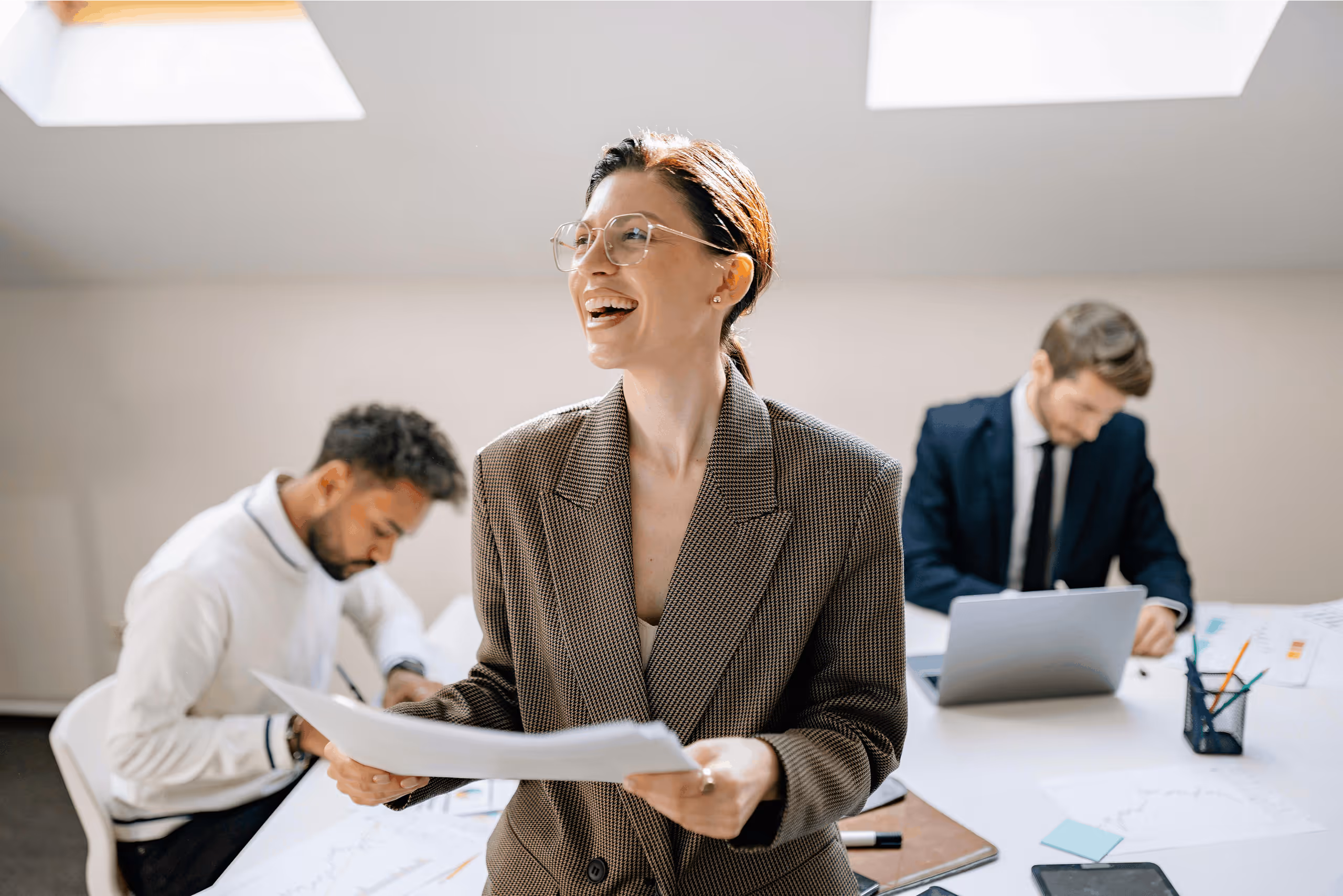 Smiling businesswoman holding papers, with two men working at laptops in bright office.