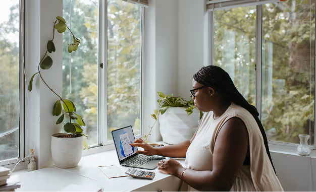 Woman using a laptop and calculator at a bright desk near windows with potted plants.