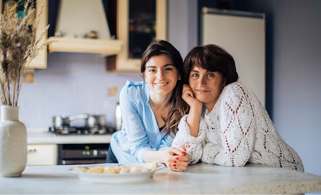 Two women smiling and leaning on a kitchen counter with a plate of food in front of them.