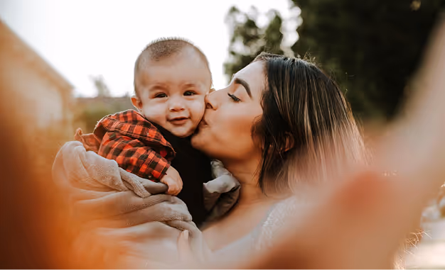 Woman kissing a smiling baby wrapped in a blanket outdoors with blurred foreground and background.