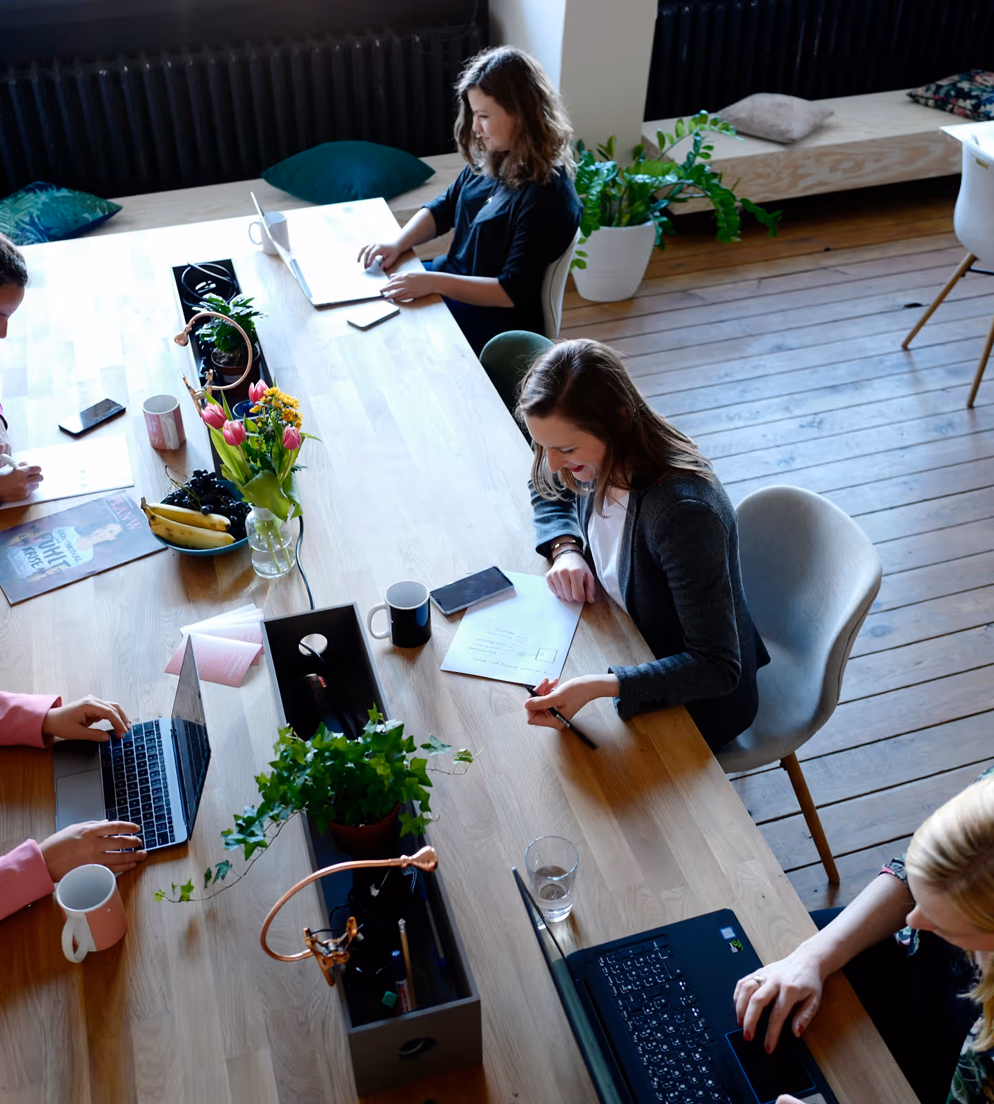 Four people working around a wooden table with laptops, papers, mugs, and plants in a bright room.