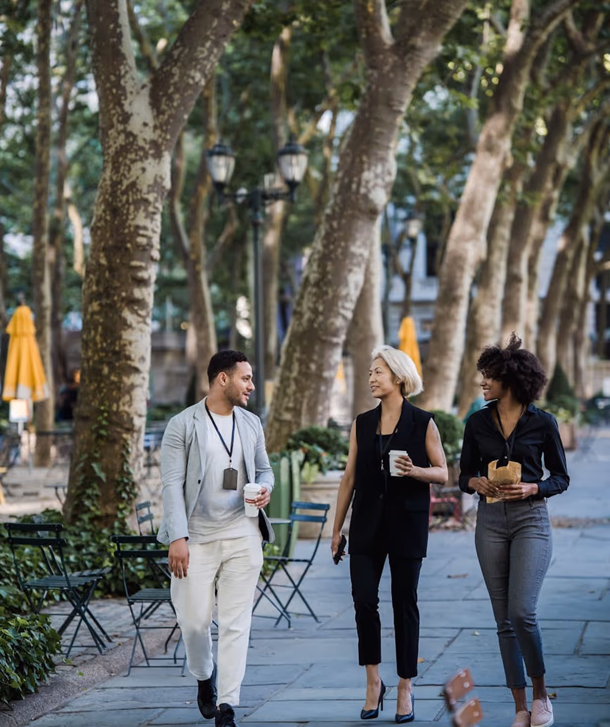 Three diverse colleagues walking and talking outside in a tree-lined urban park with drinks and snacks.
