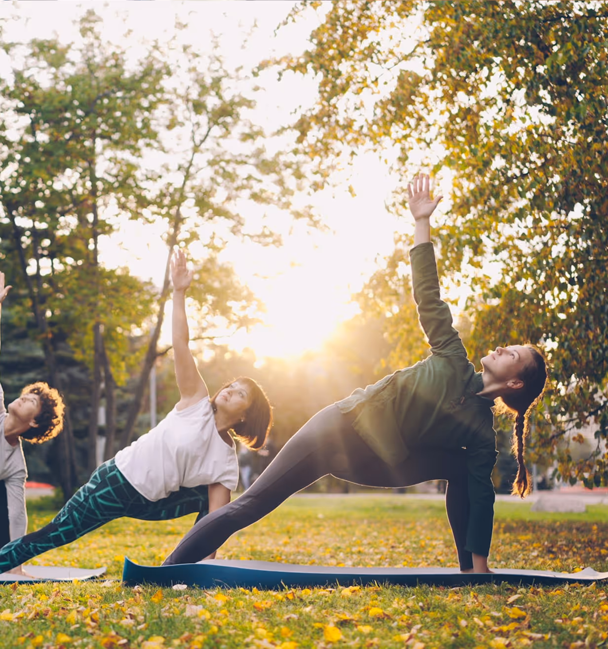 Three people practicing yoga outdoors on mats, stretching side poses with arms raised toward the sun.