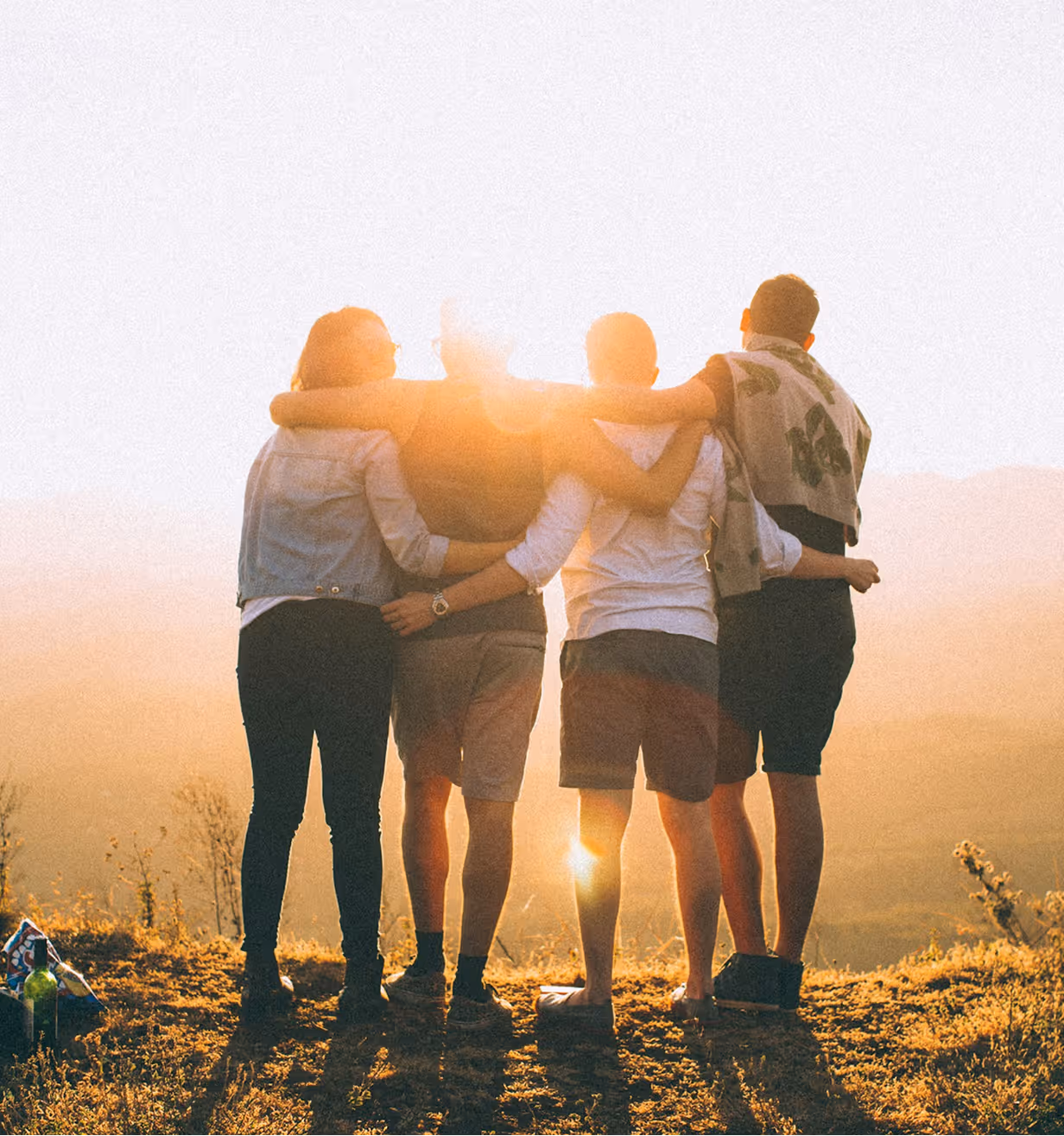 Four people stand arm in arm on a hill at sunset, facing away toward the horizon.