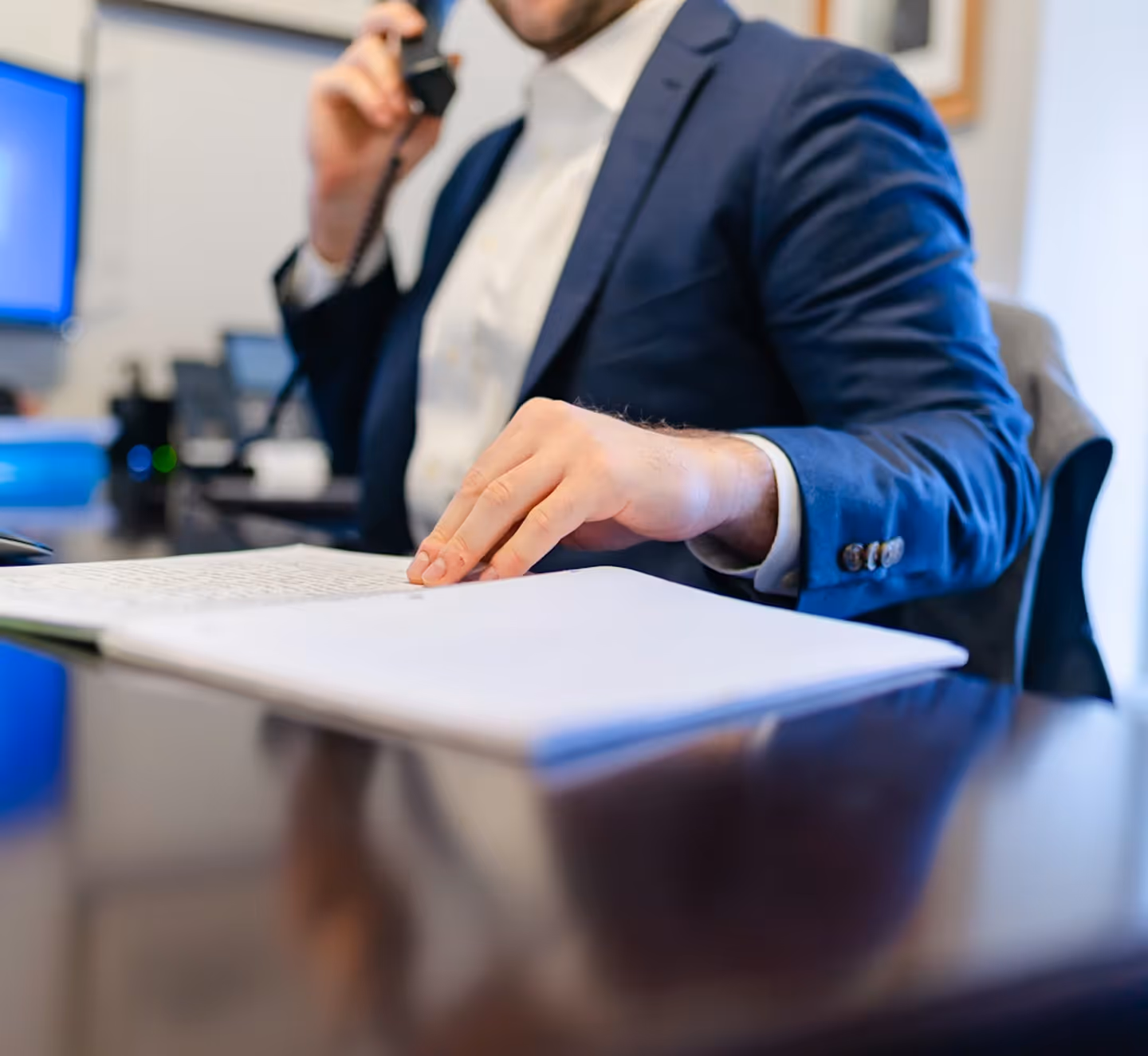 Man in a blue suit talking on phone while reviewing documents at a desk with computer screens nearby.
