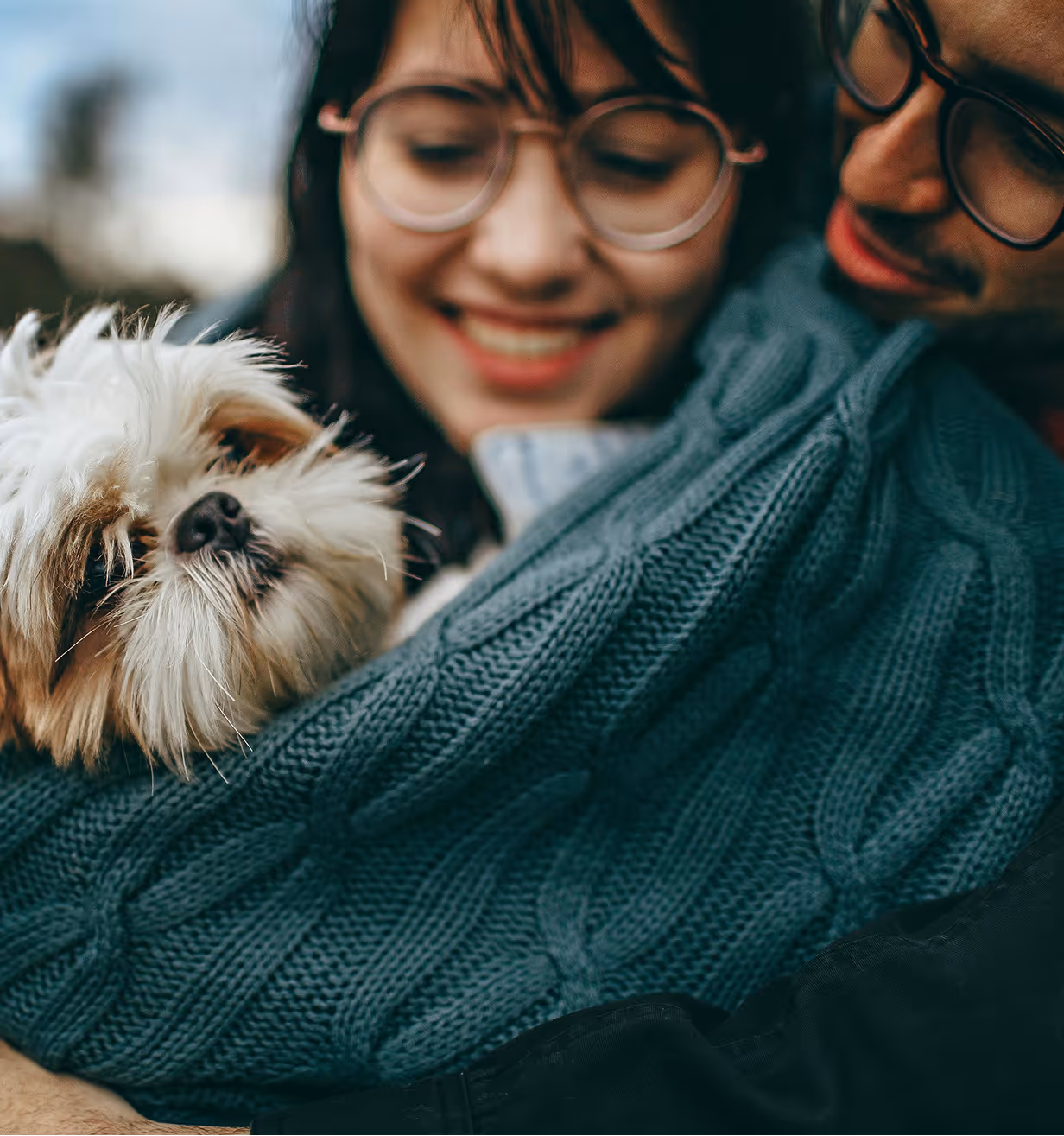 A couple smiling and holding a small fluffy white and brown dog wrapped in a blue knitted blanket.