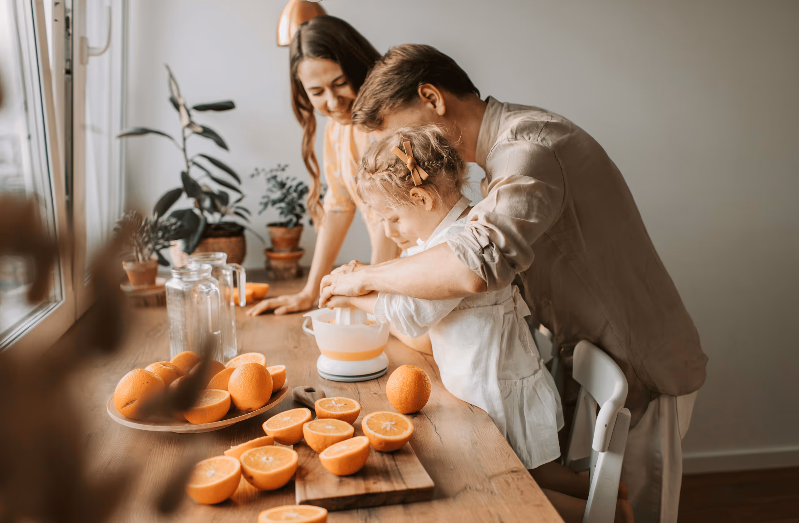 A man and woman help a girl juice oranges at a kitchen table with sliced oranges and plants nearby.