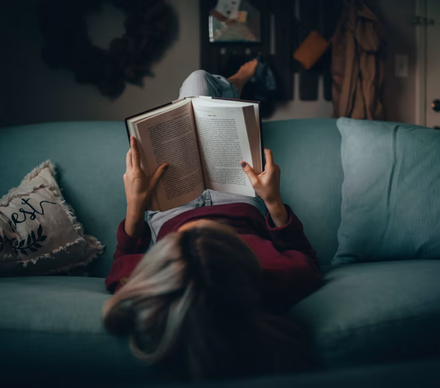Person lying on a green couch reading a book indoors with legs crossed and a pillow beside them.