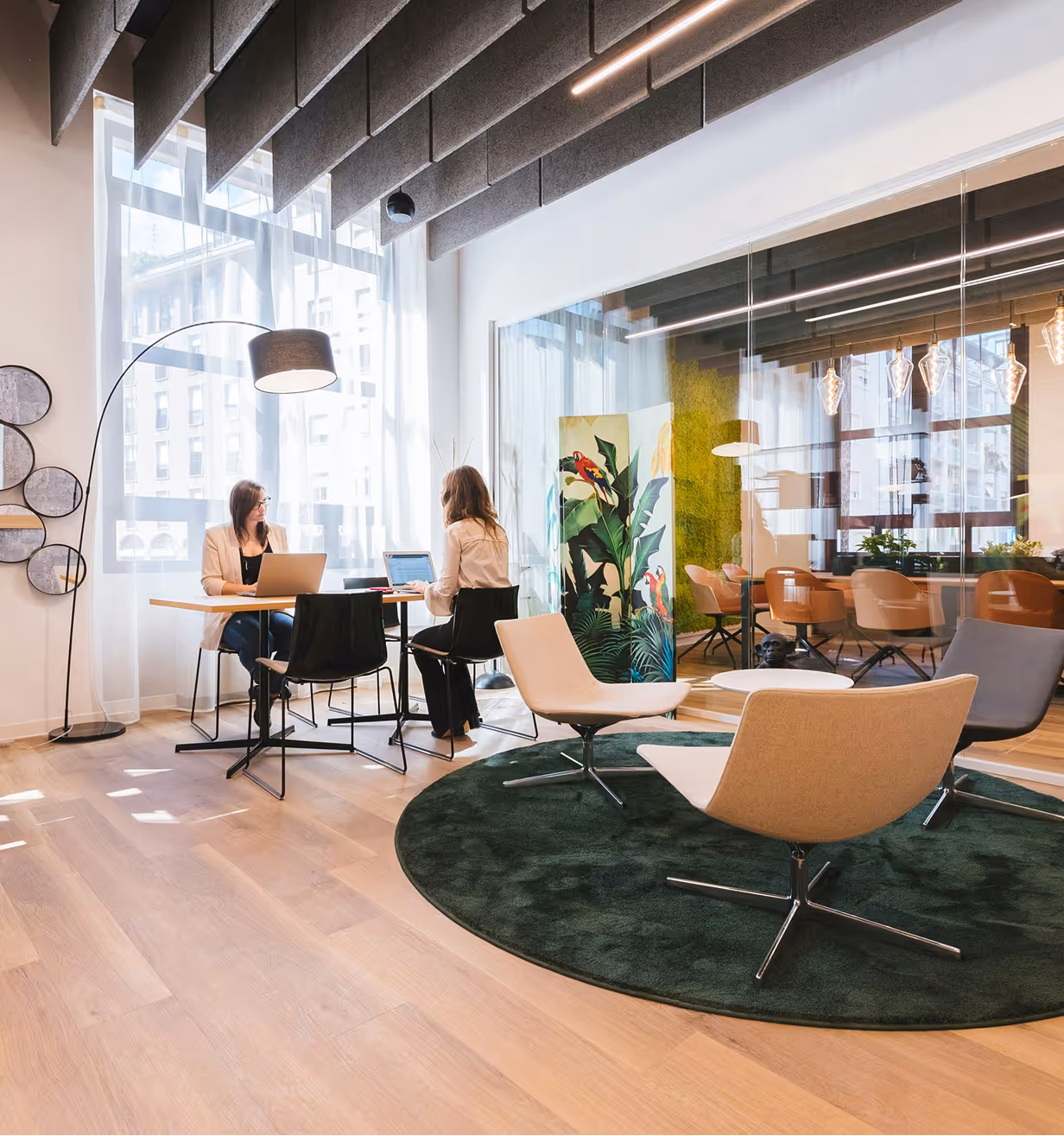 Modern office space with two women working at a table and minimalist lounge chairs on a green rug.