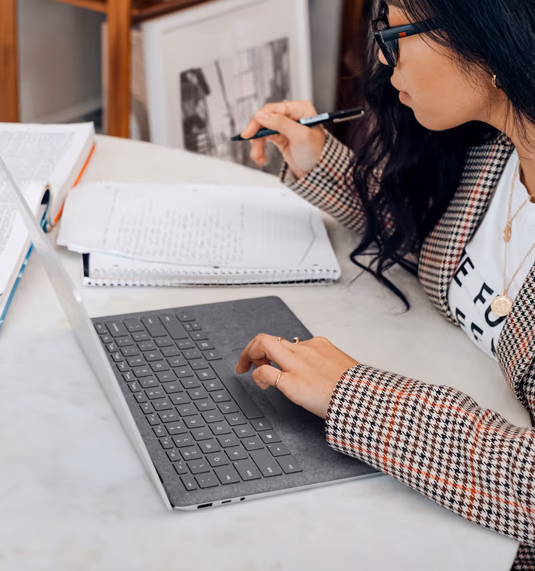 Woman with glasses working on a laptop and writing notes on paper at a desk.