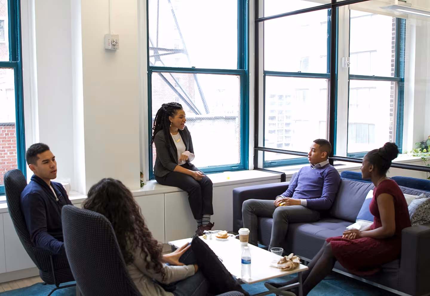 Four people in casual clothing having a discussion in a bright office lounge with large windows.