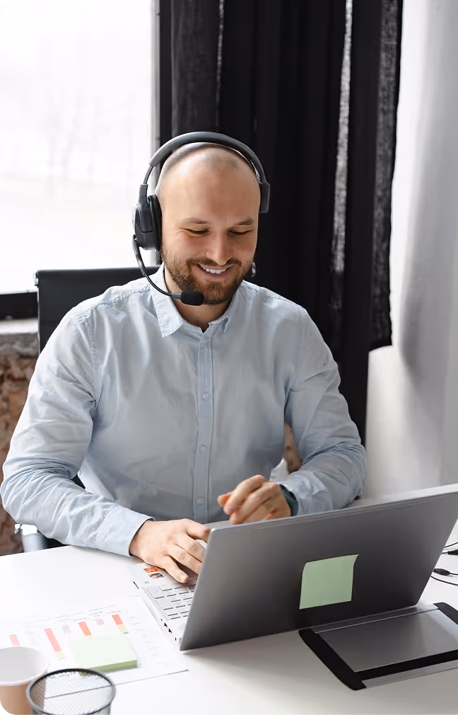 Smiling man wearing headset working on laptop at a bright office desk with charts and sticky notes.