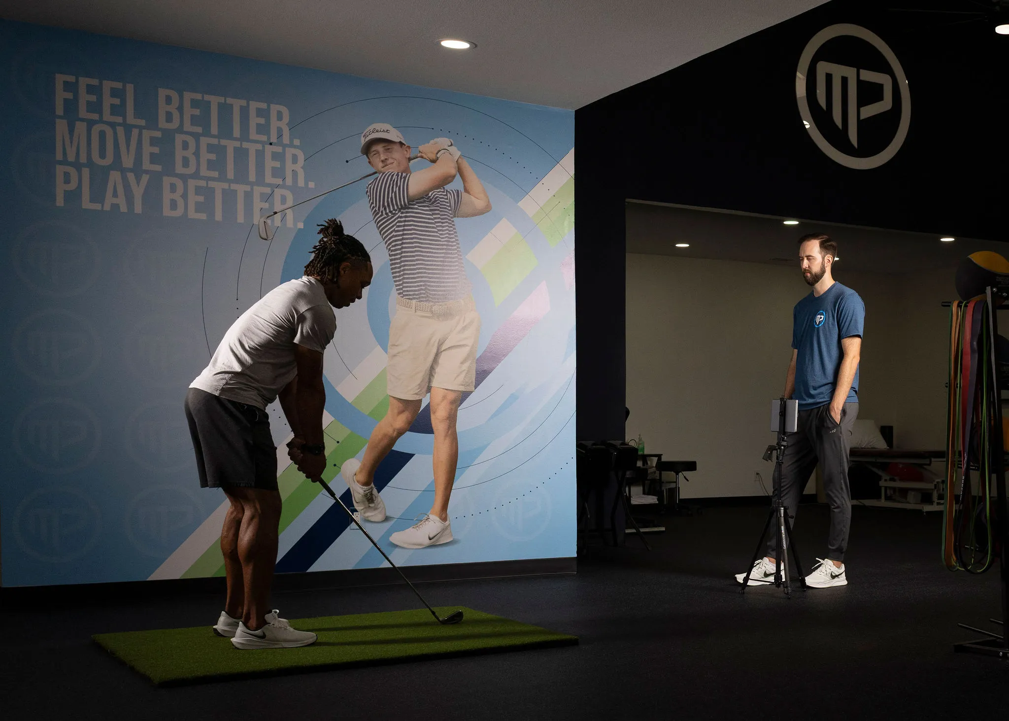 Man practicing golf swing indoors on green mat while another man observes with equipment.