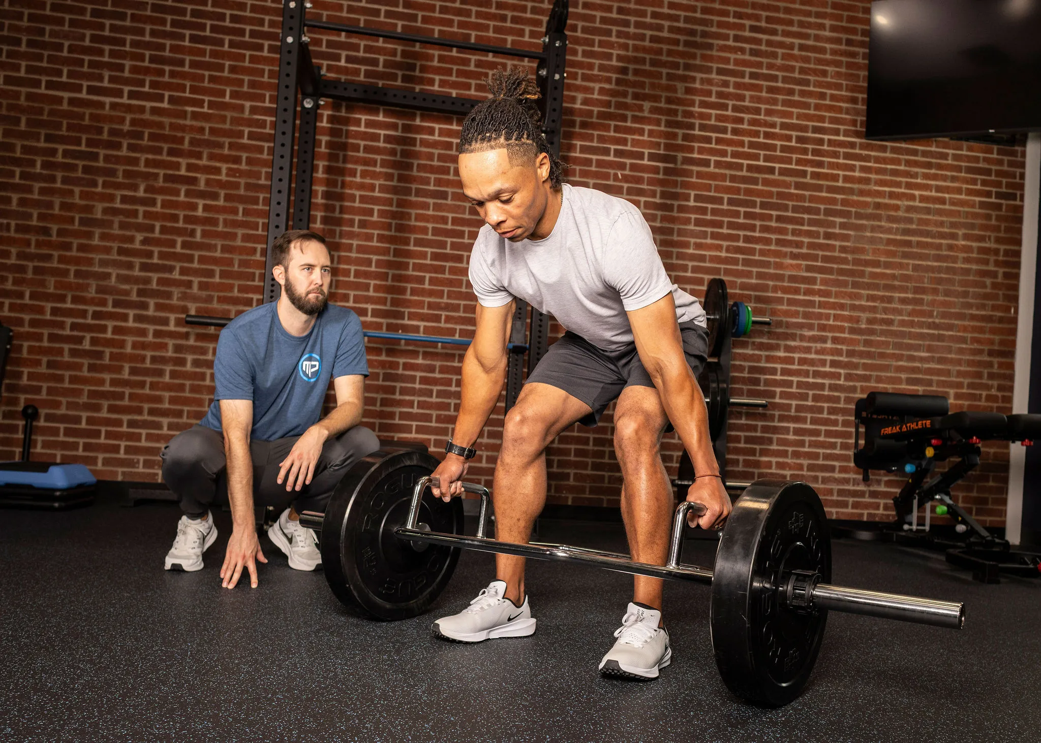 Man lifting a trap bar weight in a gym, coached by a squatting man in a blue shirt and white sneakers.
