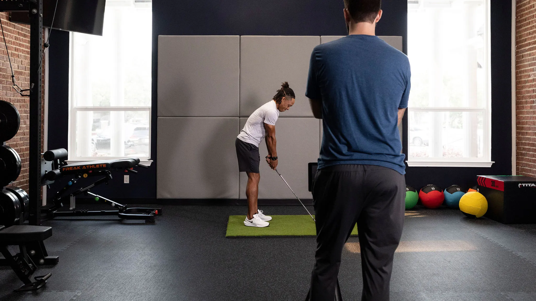 Man practicing golf putting on green mat indoors with another man observing in gym setting.
