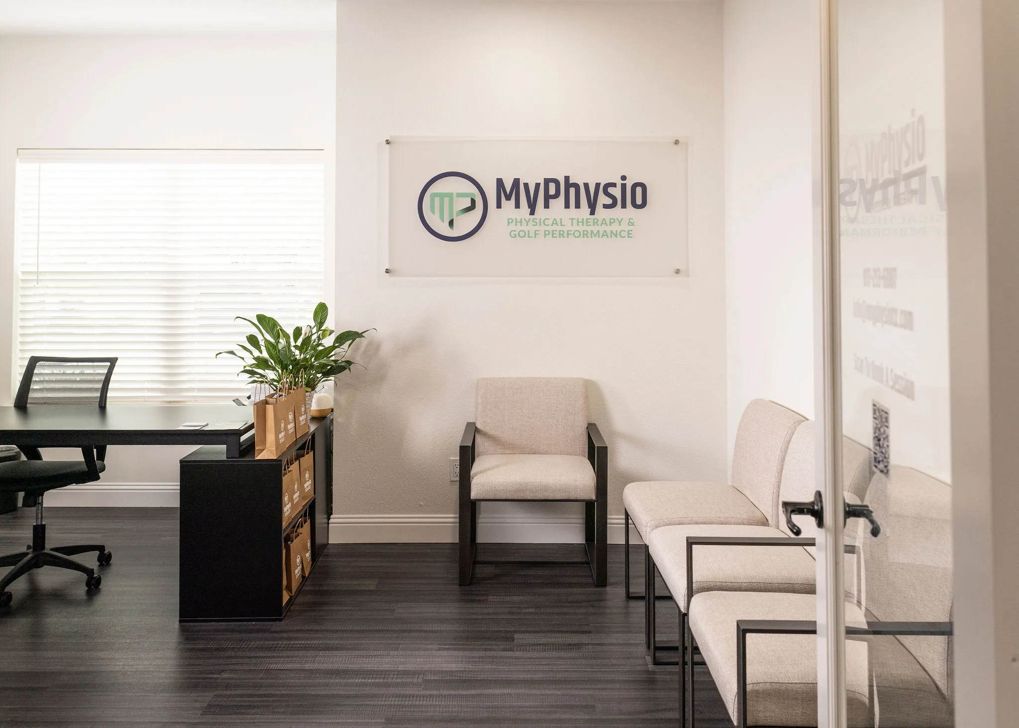 Modern physical therapy office waiting area with beige chairs, desk, plant, and MyPhysio sign on wall.