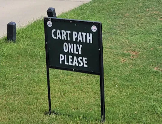 Sign reading "CART PATH ONLY PLEASE" on a grassy area by a paved path, with a calm outdoor setting.