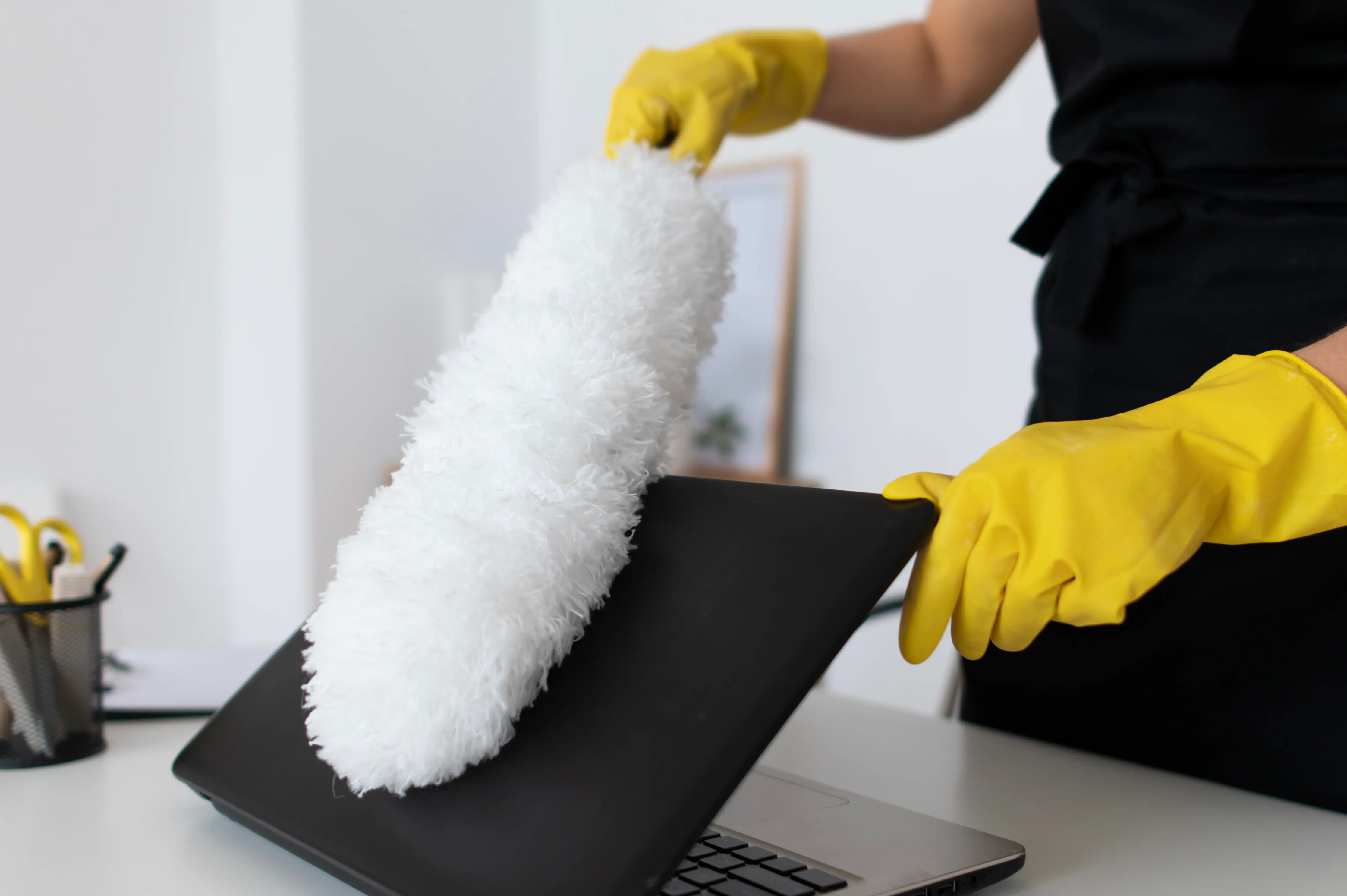 Person wearing yellow gloves cleaning a black laptop with a white fluffy duster.