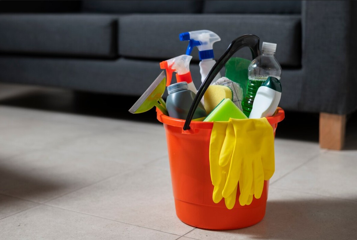 Orange bucket filled with cleaning supplies including spray bottles, sponges, and yellow rubber gloves.