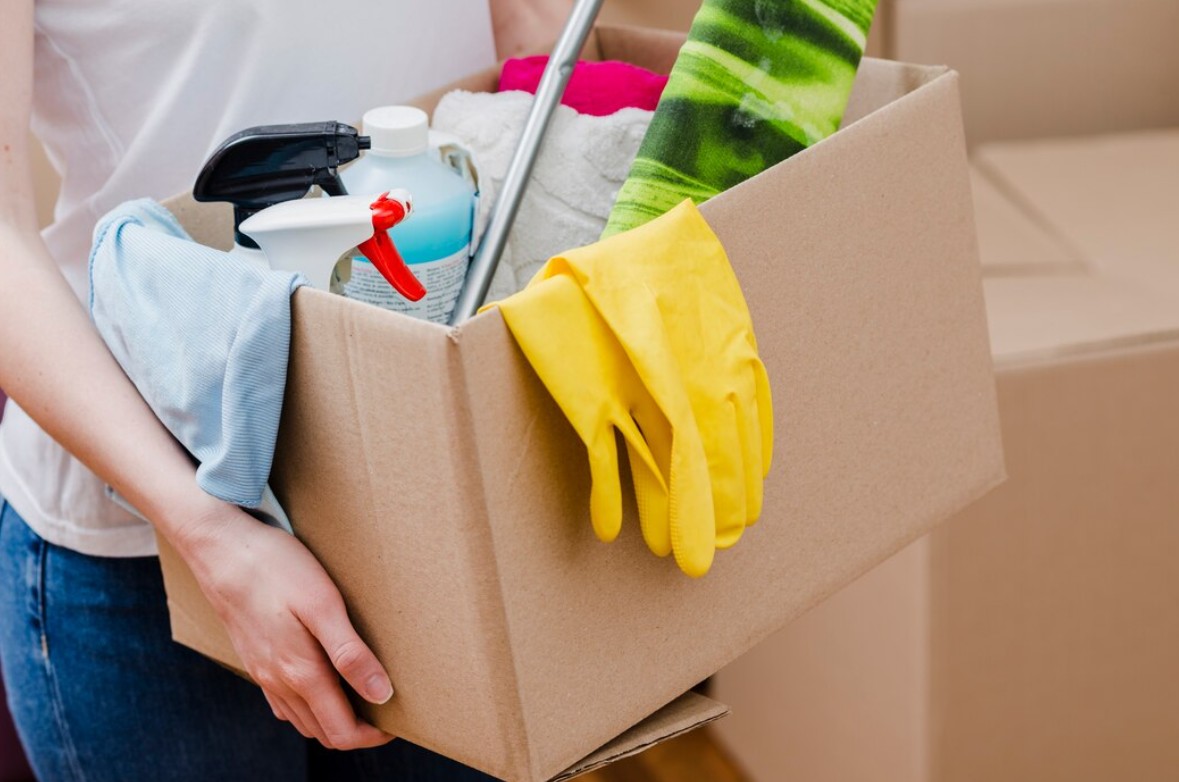 Person holding a box filled with cleaning supplies including gloves, spray bottles, and cloths.