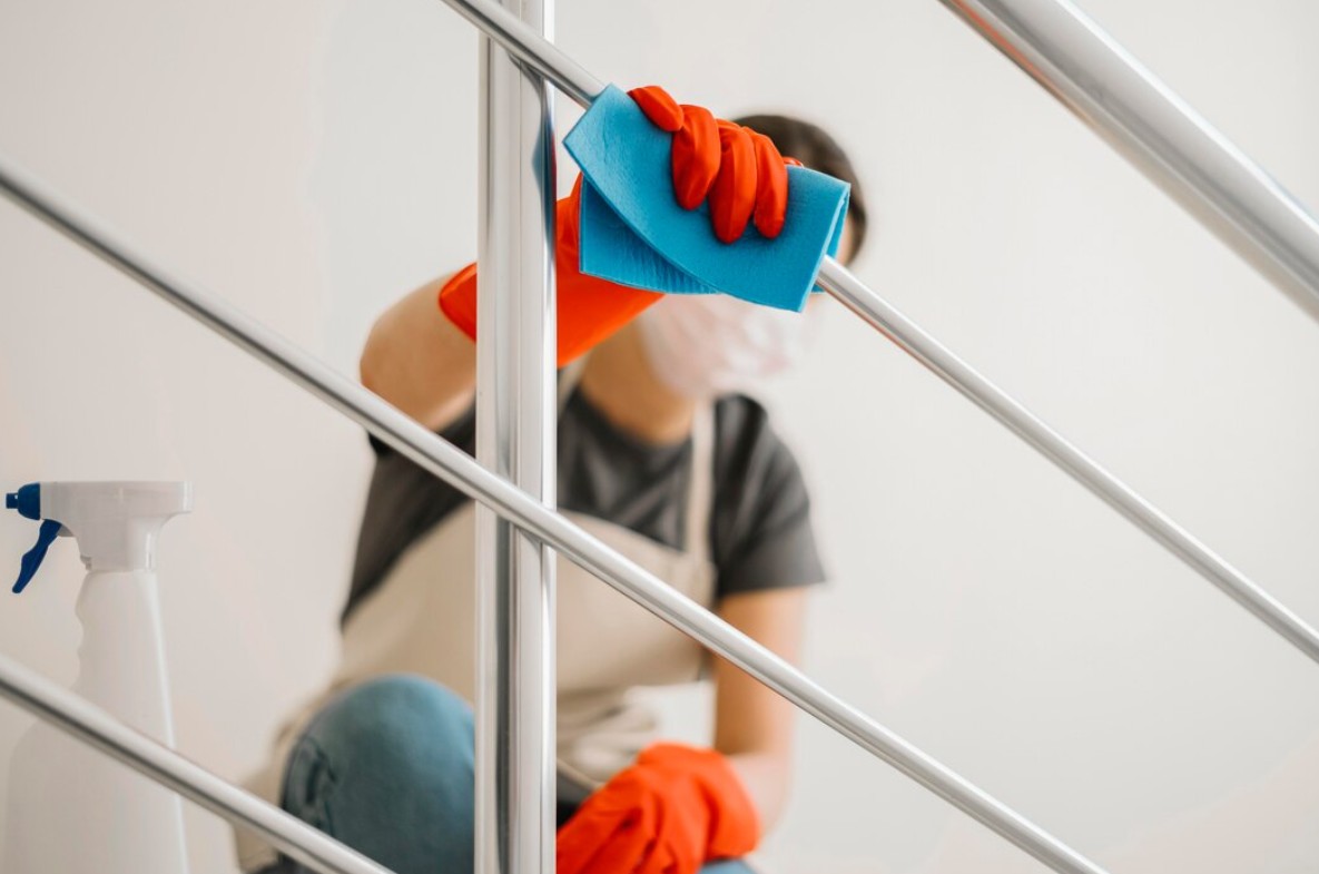 Person wearing orange gloves cleaning a silver metal railing with a blue cloth and spray bottle nearby.