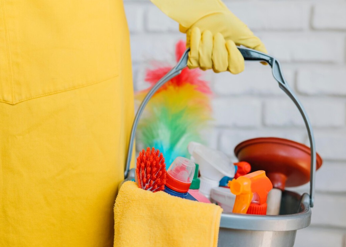Person wearing yellow gloves and apron holding bucket with cleaning supplies and tools.