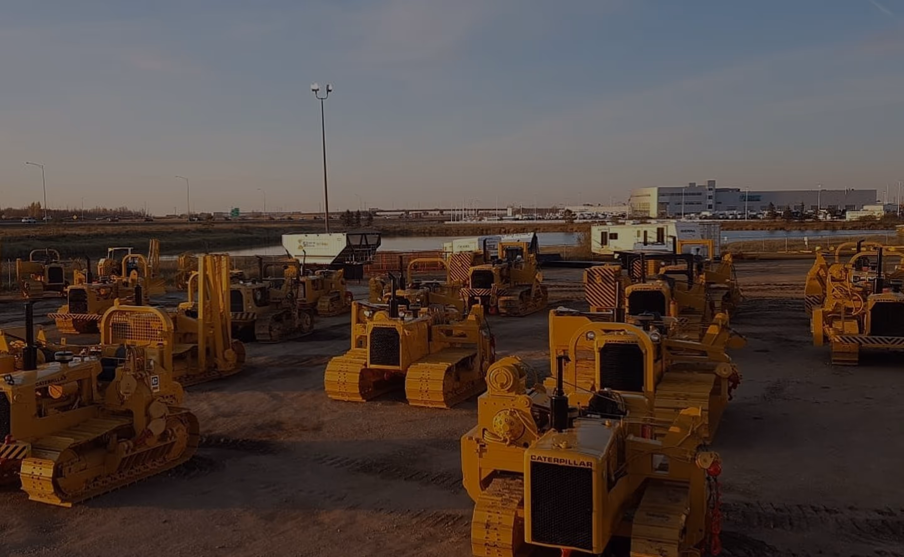 Lot filled with several yellow Caterpillar bulldozers parked on dirt ground near water and buildings.