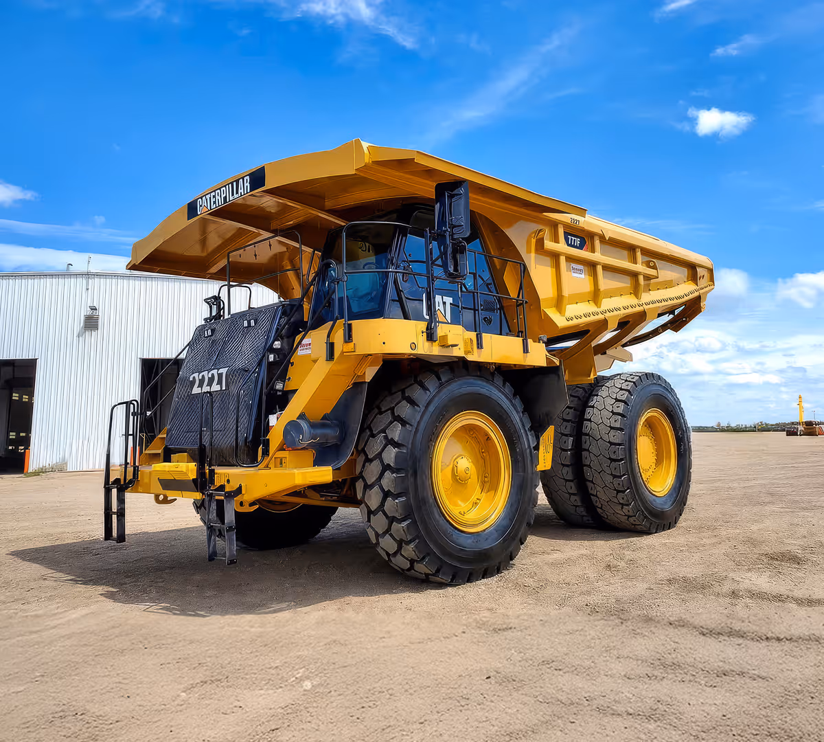 Large yellow Caterpillar 777F dump truck parked on dirt near a white metal building under blue sky.