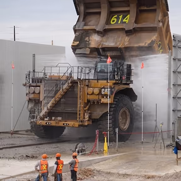 Large CAT mining truck being washed with high-pressure water while three workers in safety gear observe.