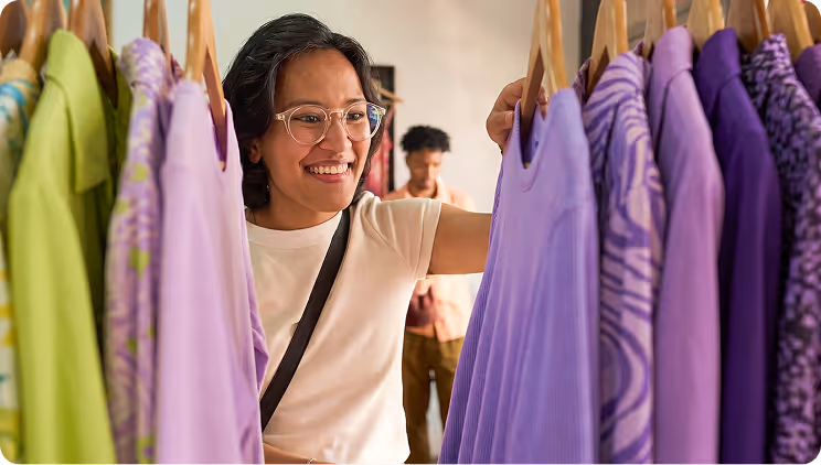 Smiling woman with glasses browsing purple and green clothes on hangers in a store.