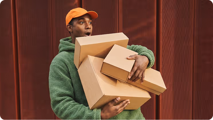 Man in green hoodie and orange cap surprised while holding multiple cardboard boxes.