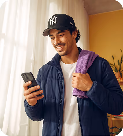 Smiling man in New York Yankees cap and jacket holding a phone and purple towel indoors.
