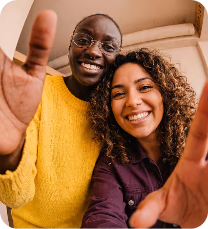 Two smiling women reaching toward the camera, one in a yellow sweater, the other with curly hair in a purple shirt.