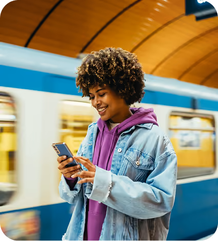 Smiling woman in purple hoodie and denim jacket using smartphone at a subway station with a blue train.