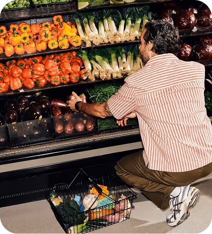 Man crouching and selecting vegetables from a supermarket shelf with a basket on the floor.