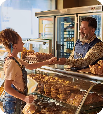 Child paying a smiling bakery worker over a glass pastry display filled with baked goods.