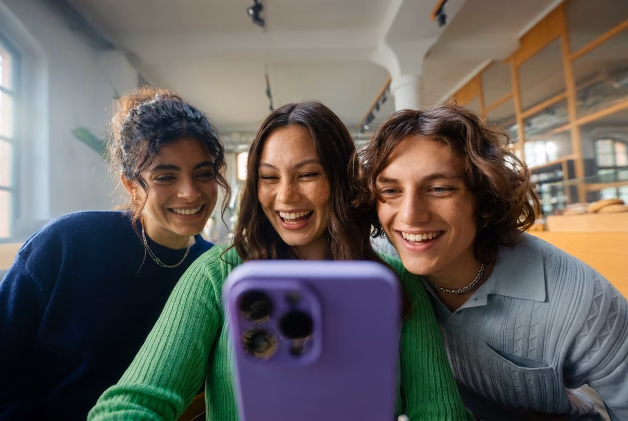 Three young adults smiling and looking at a smartphone with a purple case, sitting close together indoors with natural light and wooden interior in the background.