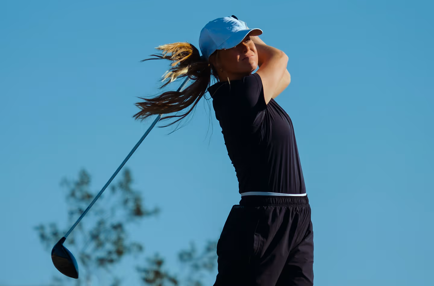 Female golfer in a dark outfit and light blue cap completing a driver swing against a bright blue sky, hair swept back by motion
