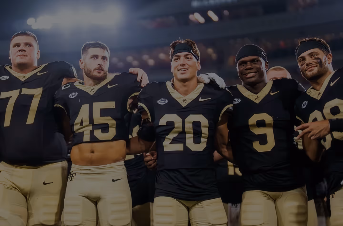 Five Wake Forest University football players in black and gold uniforms standing arm-in-arm on a stadium field under bright lights