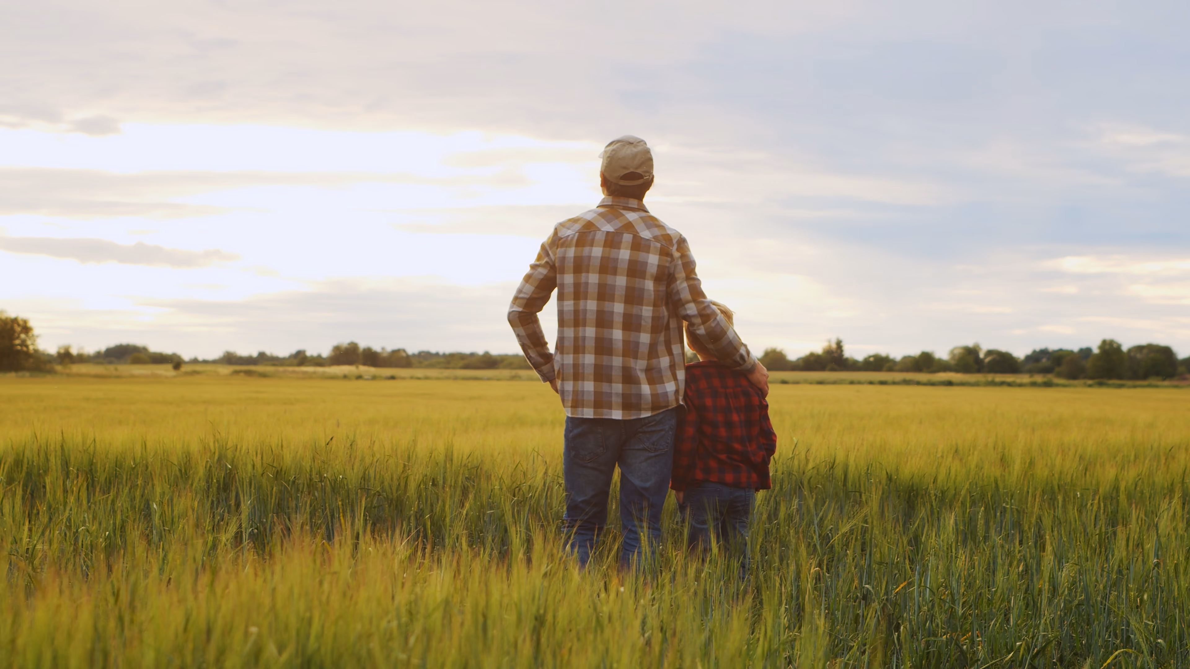 Man with arm around child standing in green field facing a cloudy sunset sky.