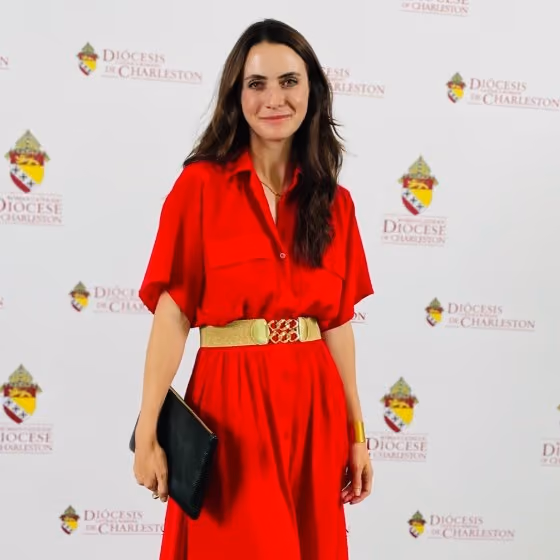 Woman with long dark hair in a red dress and gold belt holding a black clutch, standing at a Diocese of Charleston event.
